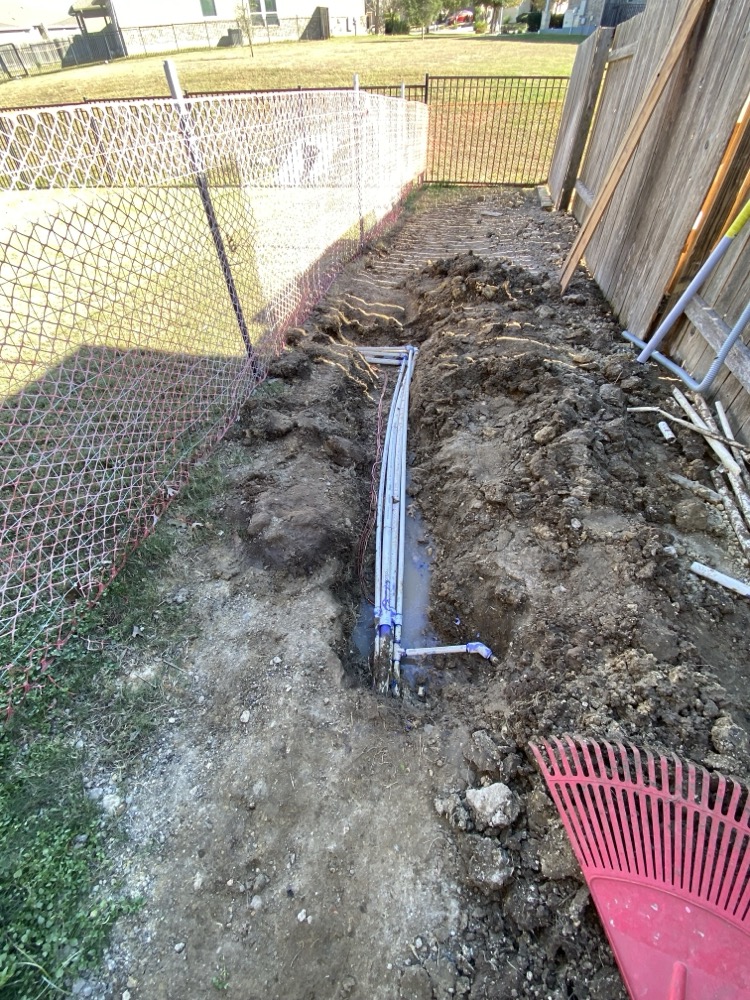 Professional irrigation installation in progress showing trenched lines with PVC main pipes and lateral connections, safety fencing visible around the work area
